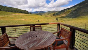 Une terrasse en bois avec une table ronde et des chaises, donnant sur une vallée où l’on aperçoit des montagnes au loin.