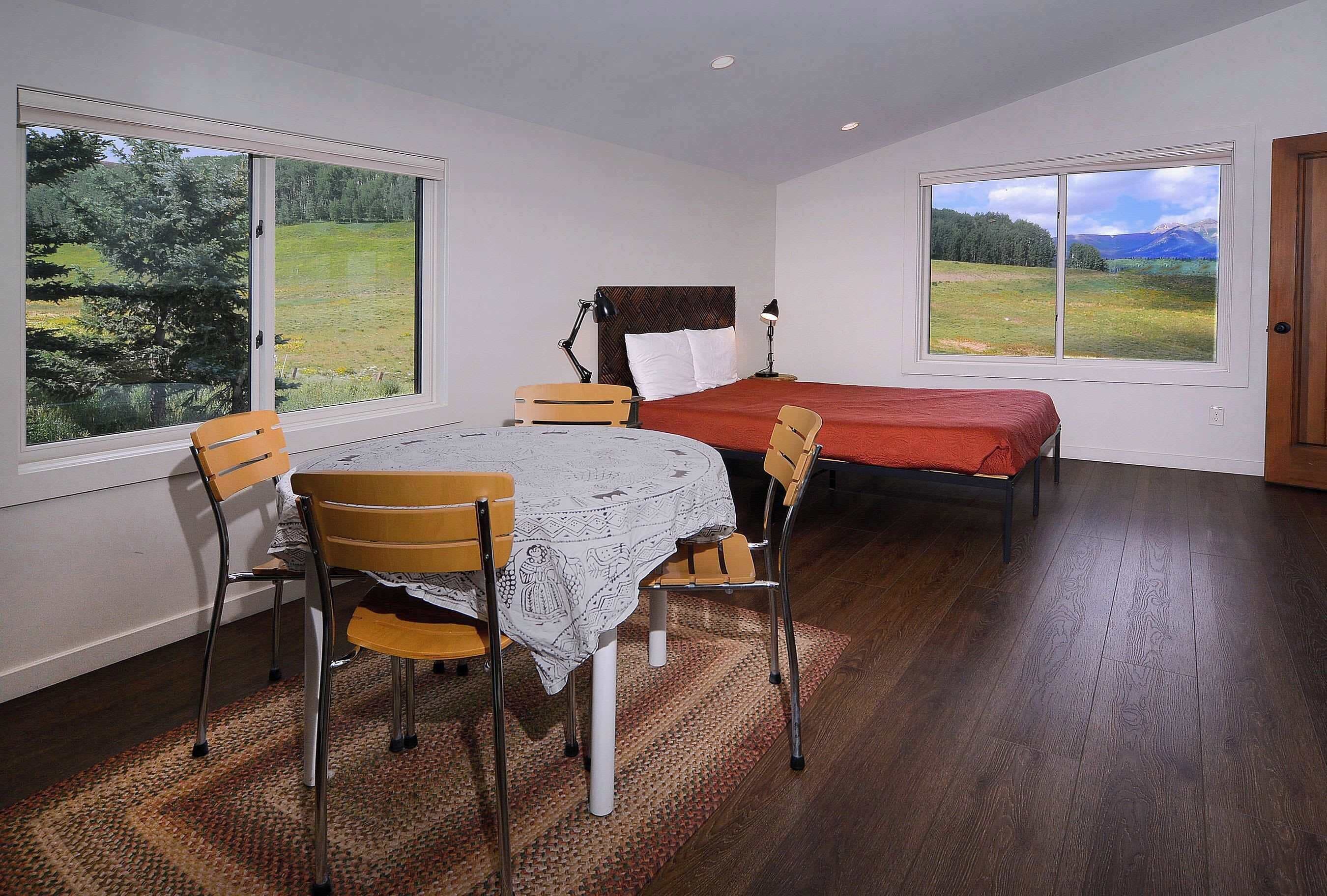 Une chambre à coucher avec un lit, une table et des chaises, et de grandes fenêtres donnant sur un champ et des montagnes.