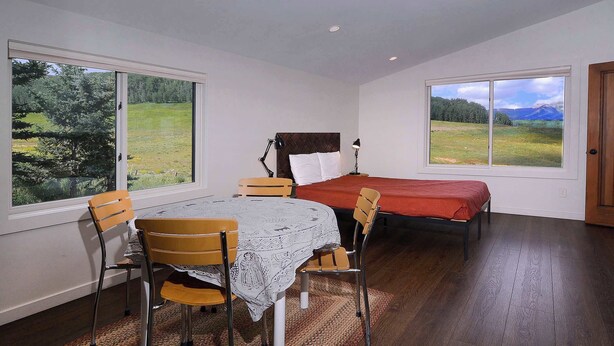 Une chambre à coucher avec un lit, une table et des chaises, et de grandes fenêtres donnant sur un champ et des montagnes.