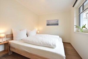 A neatly made bed with white bedding, a nightstand with a lamp, a framed picture on the wall, and a window with a plant.