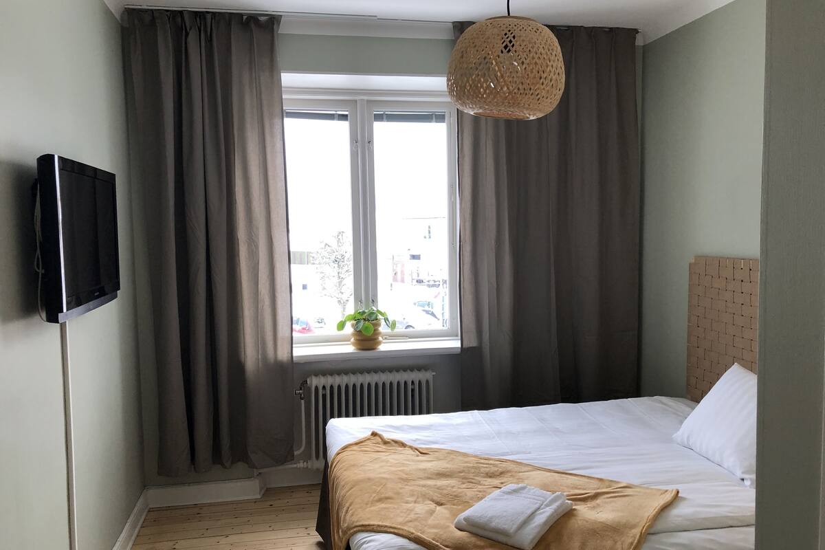 Un dormitorio con cama, televisor, ventana con cortinas y lámpara colgante.
