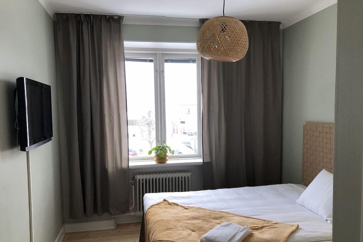 Ein Hotelzimmer mit Bett, Fenster mit Vorhängen, einer Pendelleuchte und einem Heizkörper.