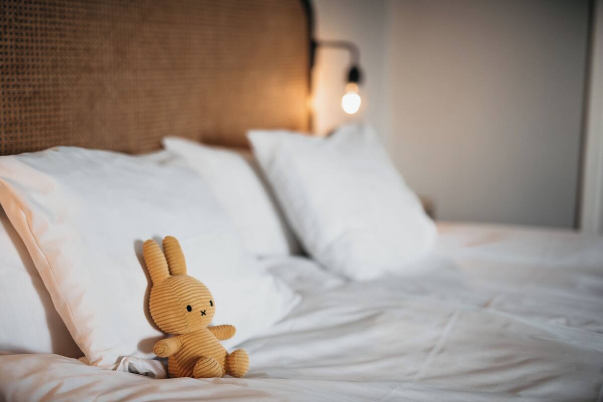 Una cama bien hecha con sábanas blancas, un pequeño conejo de madera y una lámpara de pared.