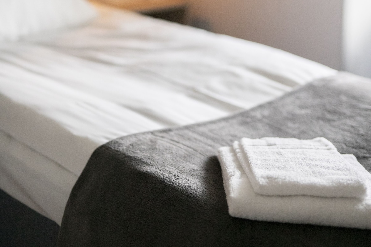 Una cama bien tendida con sábana blanca y una toalla blanca doblada sobre una mesita de noche de madera oscura.