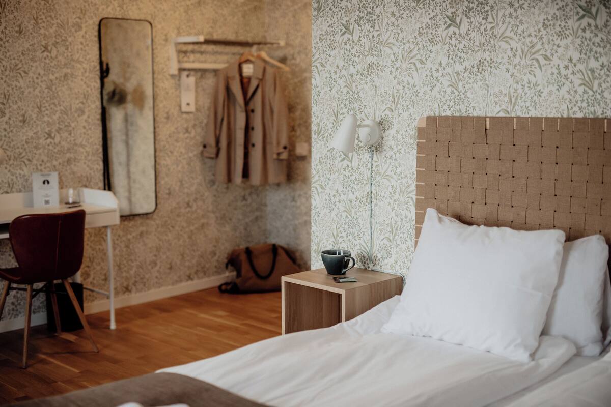 Un dormitorio con cabecera de madera, una mesita de noche con una taza, una silla, un escritorio y un perchero.