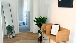 A modern, well-lit hallway with a wooden floor, a round woven rug, a wooden bench with a framed menu, a large mirror, and a potted plant. - Glück Auf Appartement Lösortstraße (Duisburg)