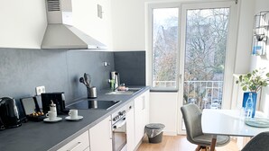 A modern kitchen with a dining area, featuring a large window, wooden flooring, and contemporary appliances. - Glück Auf Appartement Lösortstraße (Duisburg)