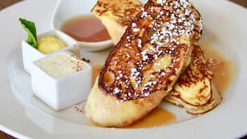 A plate of French toast with syrup and powdered sugar, accompanied by two small containers of butter and a small bowl of syrup.