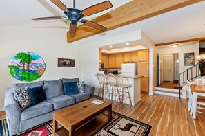 A living room with a sofa, coffee table, and a ceiling fan. There is a kitchen area with wooden cabinets and a refrigerator.
