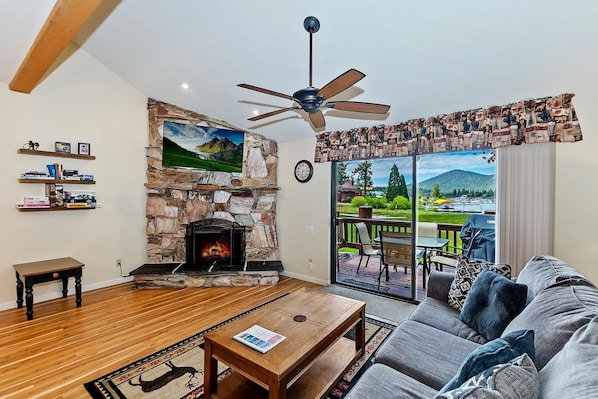 A living room with a stone fireplace, a ceiling fan, and a view of a patio with outdoor furniture. - Amazing 3BR/2BA Lakefront Condo, WiFi, Outdoor Heated Spa, FREE Kayak/Bicycle Rental! (Big Bear Lake)