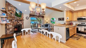 Une cuisine moderne avec une cheminée en pierre, des armoires en bois et un coin repas comprenant une grande table en bois et des chaises blanches.