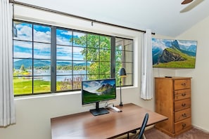 A room with a desk, a computer monitor, a lamp, a chair, a wooden dresser, and a large window with a view of a landscape. - Amazing 3BR/2BA Lakefront Condo, WiFi, Outdoor Heated Spa, FREE Kayak/Bicycle Rental! (Big Bear Lake)