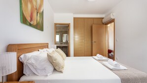 Una cama bien hecha con sábanas blancas y almohadones decorativos, una lámpara de noche y un cuadro en la pared.