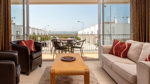 Una sala moderna con sofá, sillón y mesa de centro, un balcón con vista al mar, y un comedor con sillas y mesa.