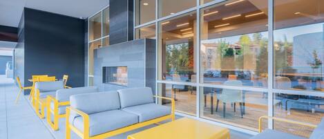A modern indoor seating area with sofas and yellow tables, large windows, and a visible outdoor view.