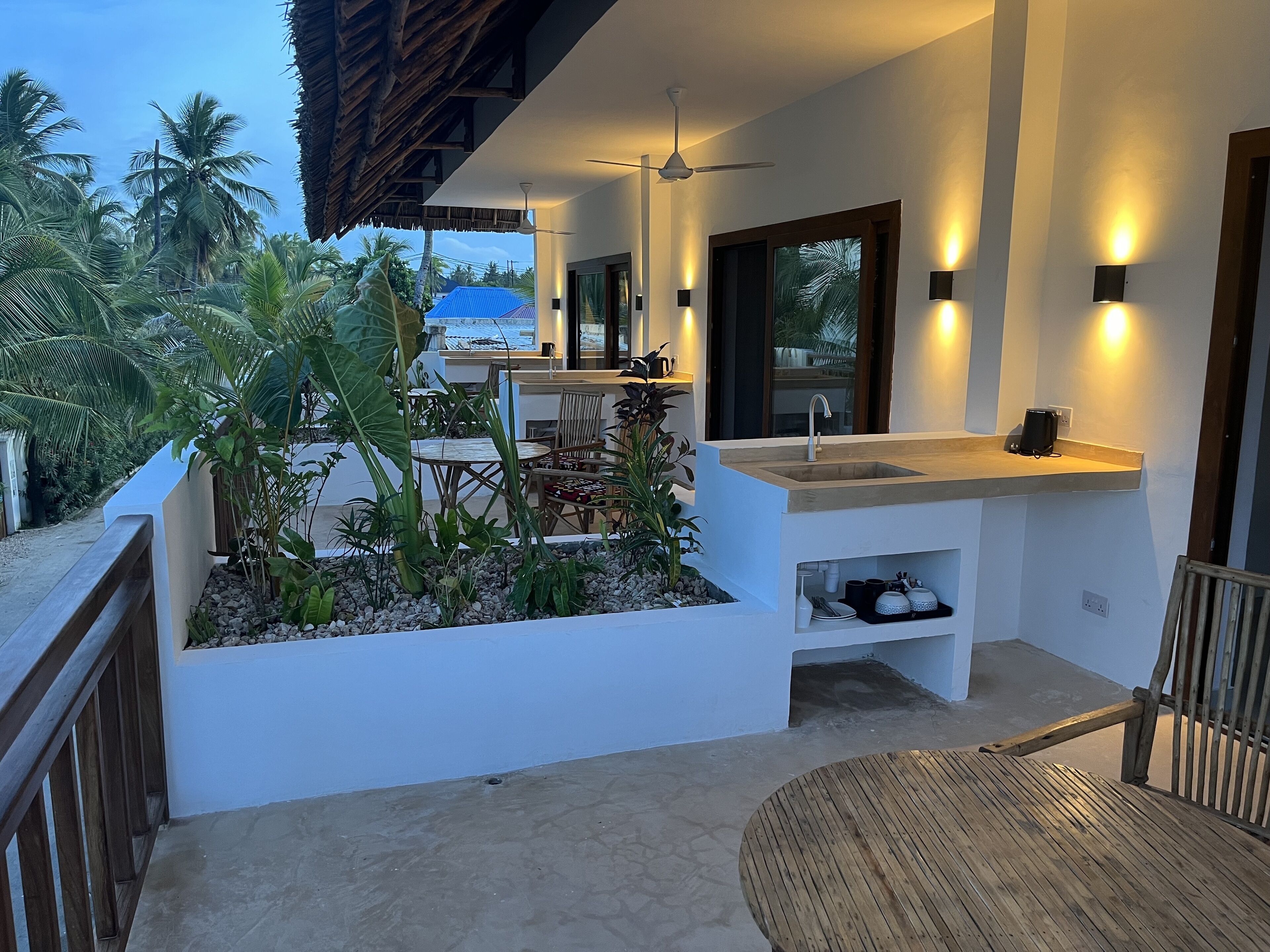 A modern outdoor kitchen area with a sink and countertop, surrounded by tropical plants and a wooden deck.