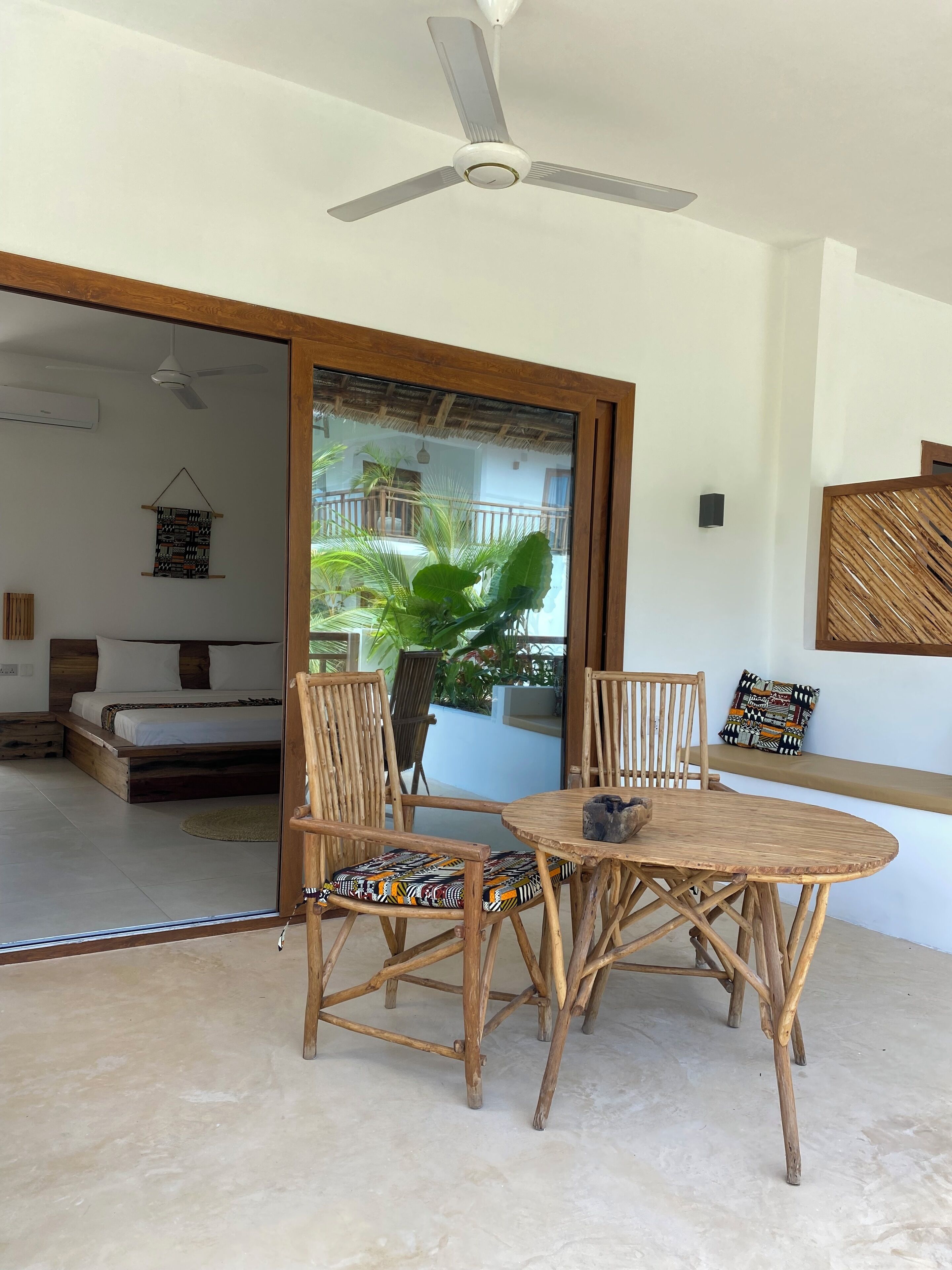 A room with a bed, a dining table with chairs, a ceiling fan, and a view of a balcony with plants.