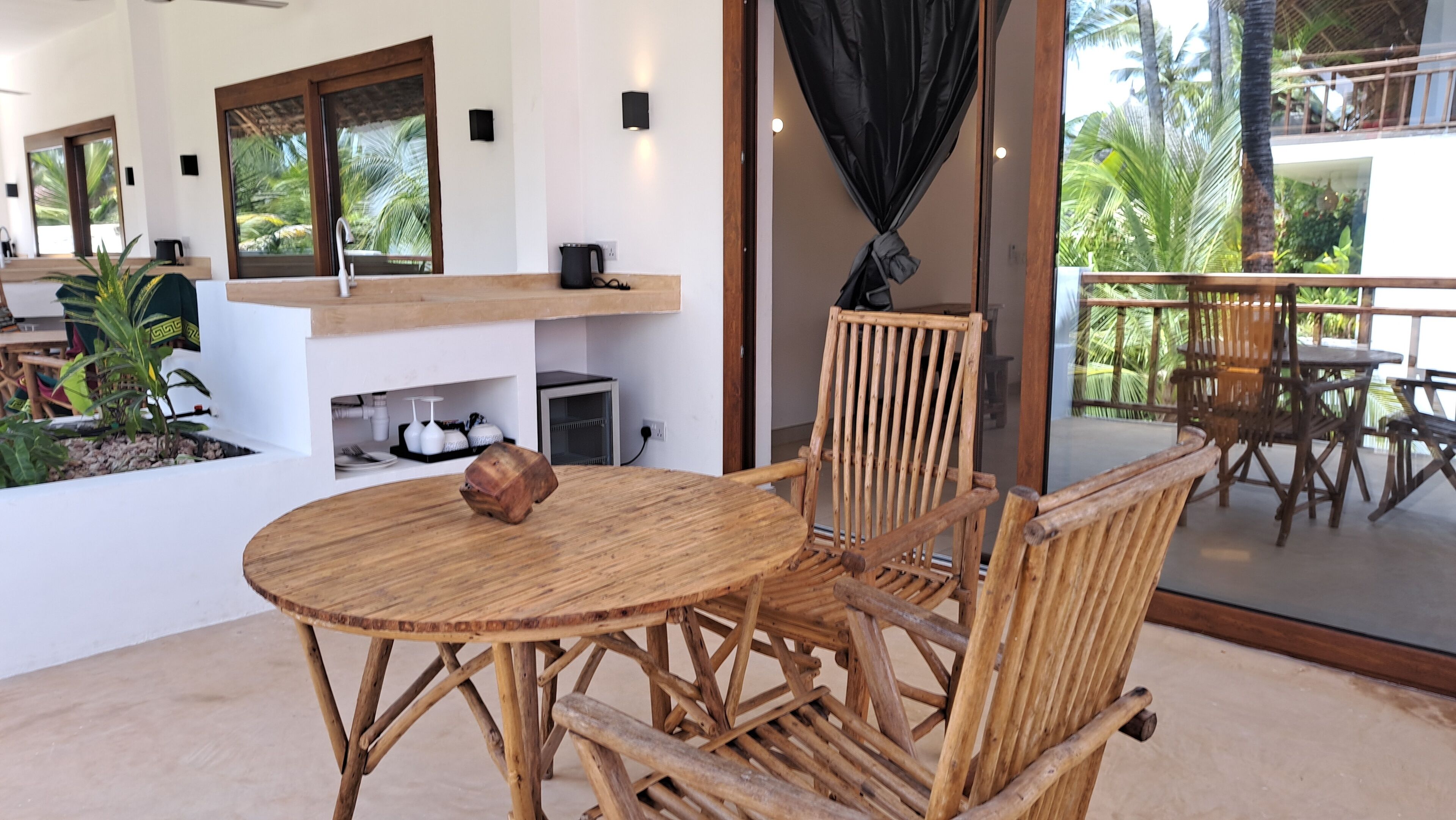A round wooden table with chairs in a room with large windows and a view of palm trees.