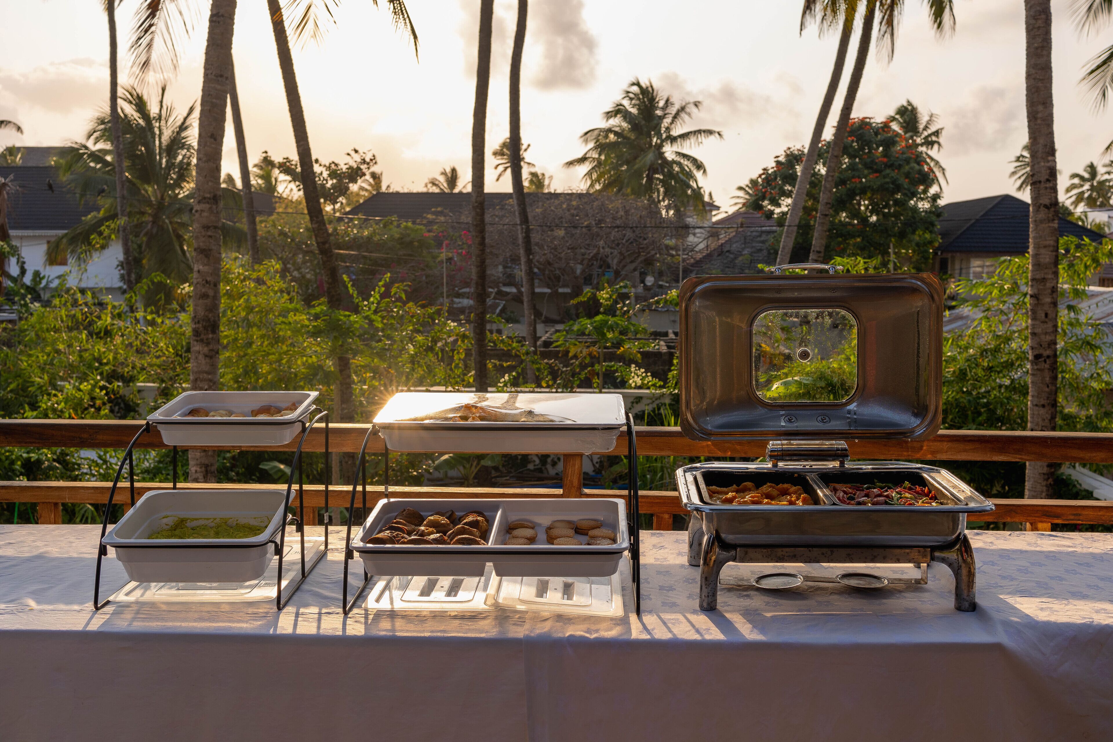 Ein reichhaltiges Buffet mit verschiedenen Speisen auf einem Tisch, umgeben von Palmen und Blick auf einen Sonnenuntergang.