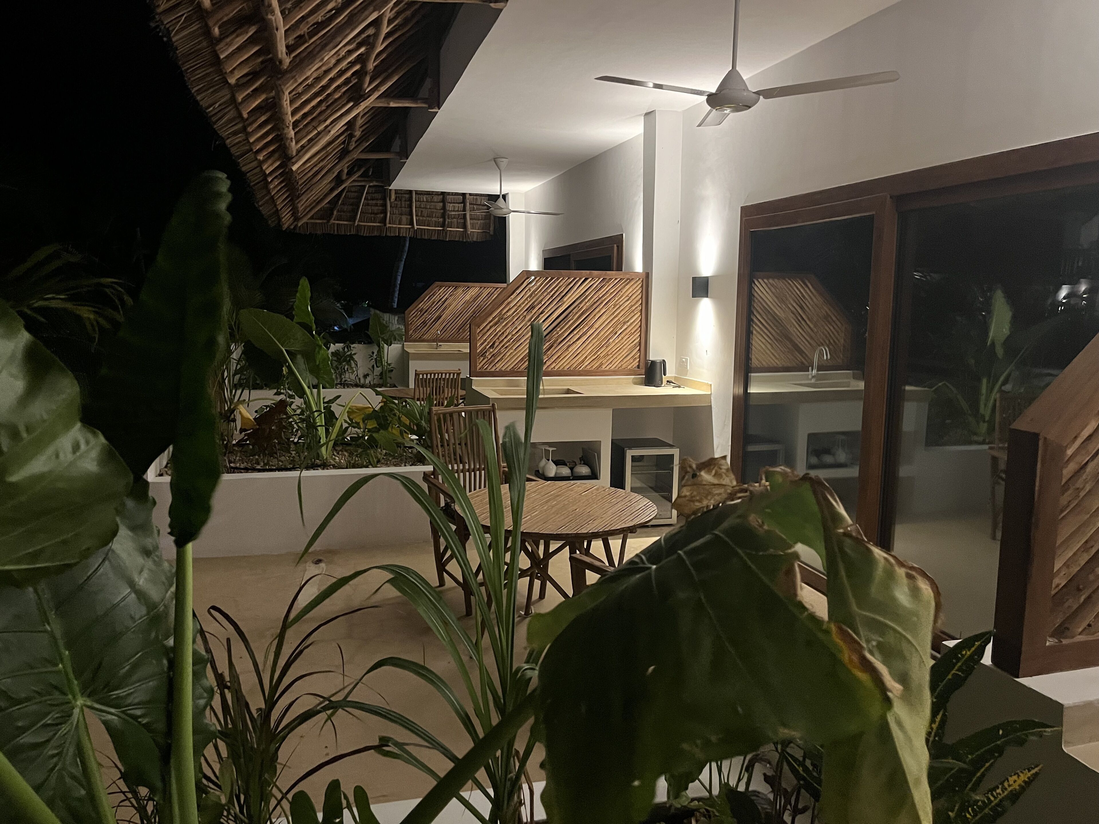 Un área de comedor al aire libre moderna con ventilador de techo, techo de paja y abundante vegetación.