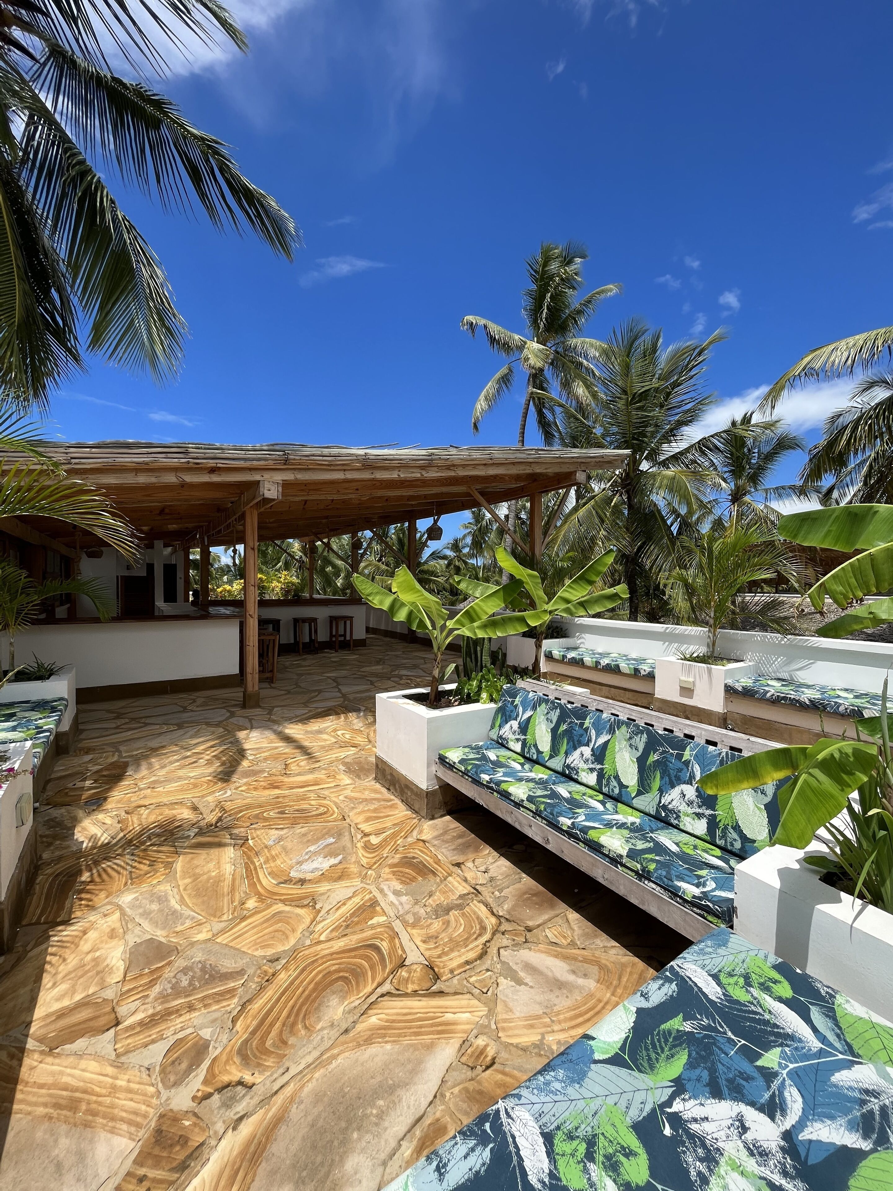 A covered outdoor seating area with stone flooring and tropical plants.