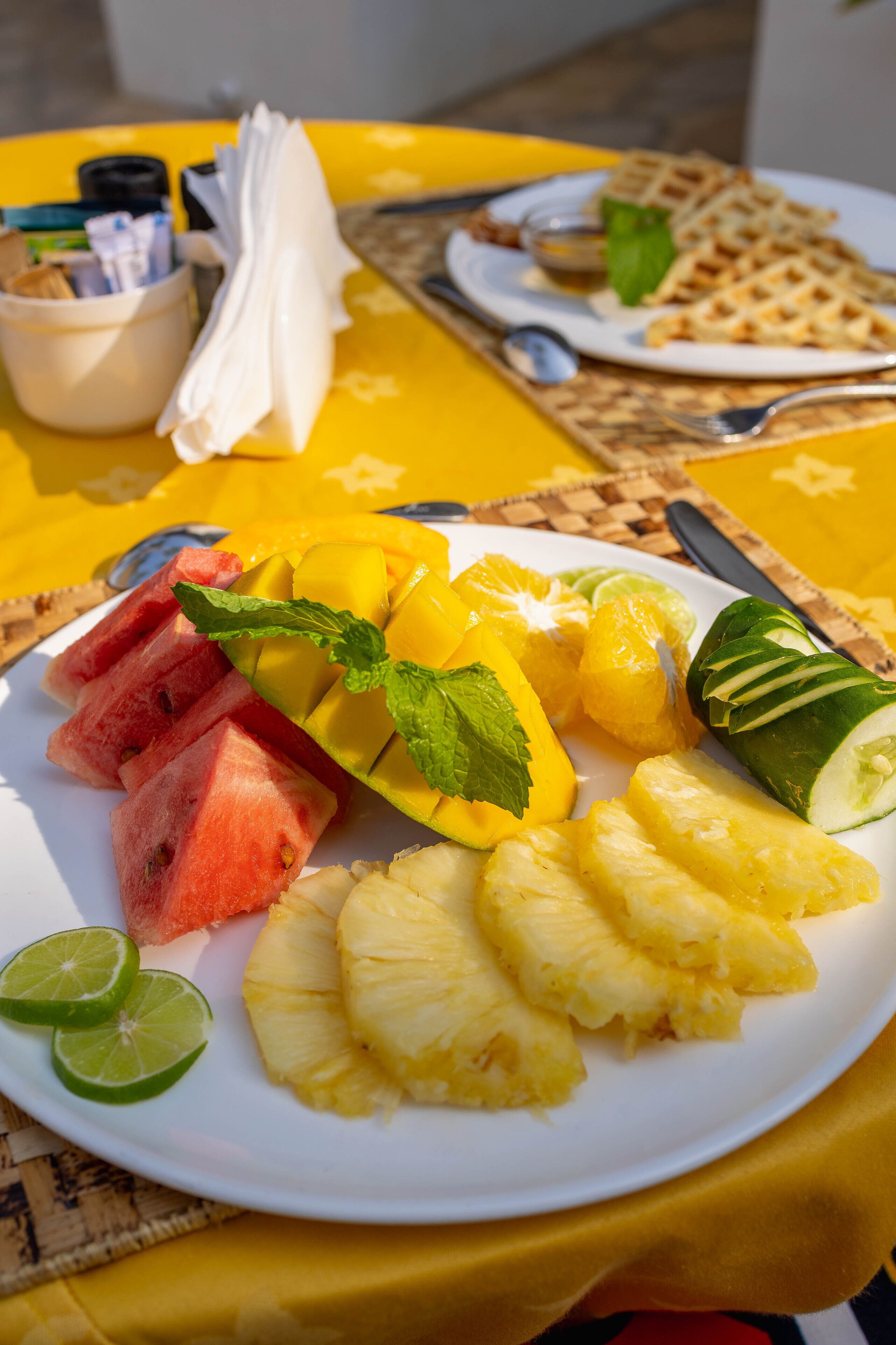 Un plato de fruta que incluye sandía, mango, piña y pepino, con una servilleta y cubiertos sobre un mantel amarillo.
