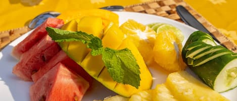 Un plato de fruta que incluye sandía, mango, piña y pepino, con una servilleta y cubiertos sobre un mantel amarillo.