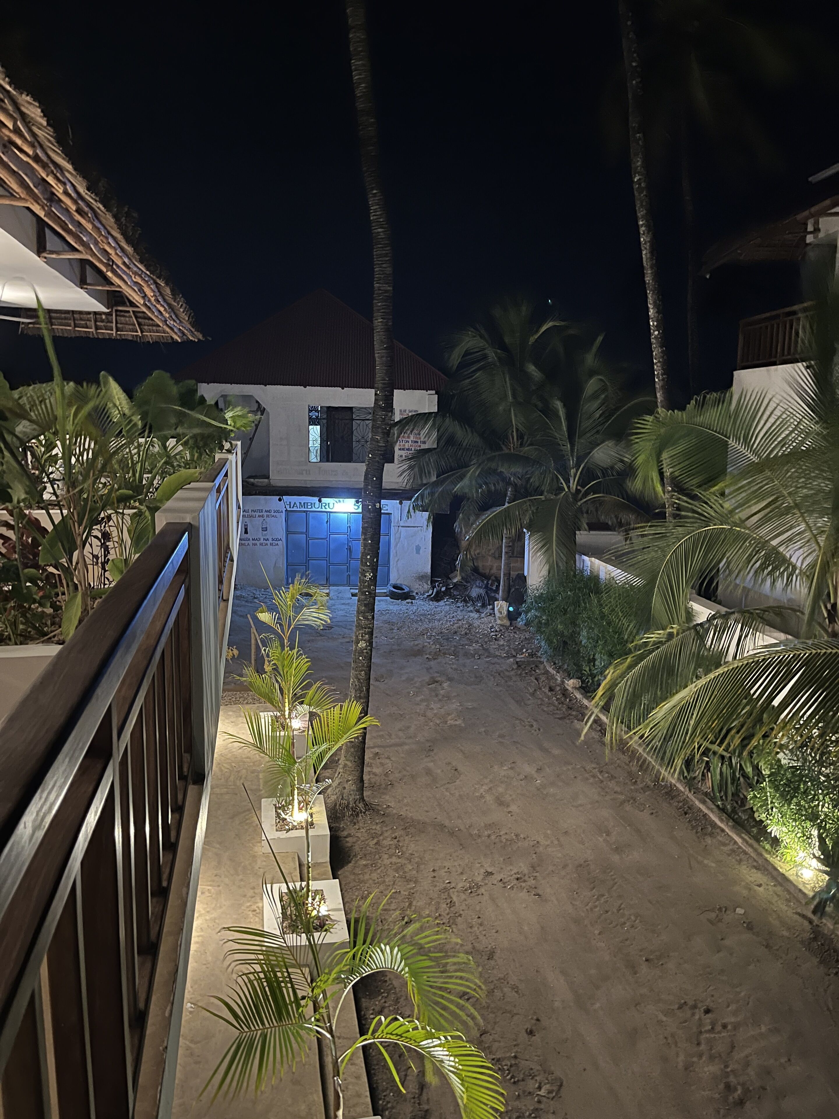 A nighttime view of a resort with a sandy pathway, palm trees, and a building with illuminated signage.