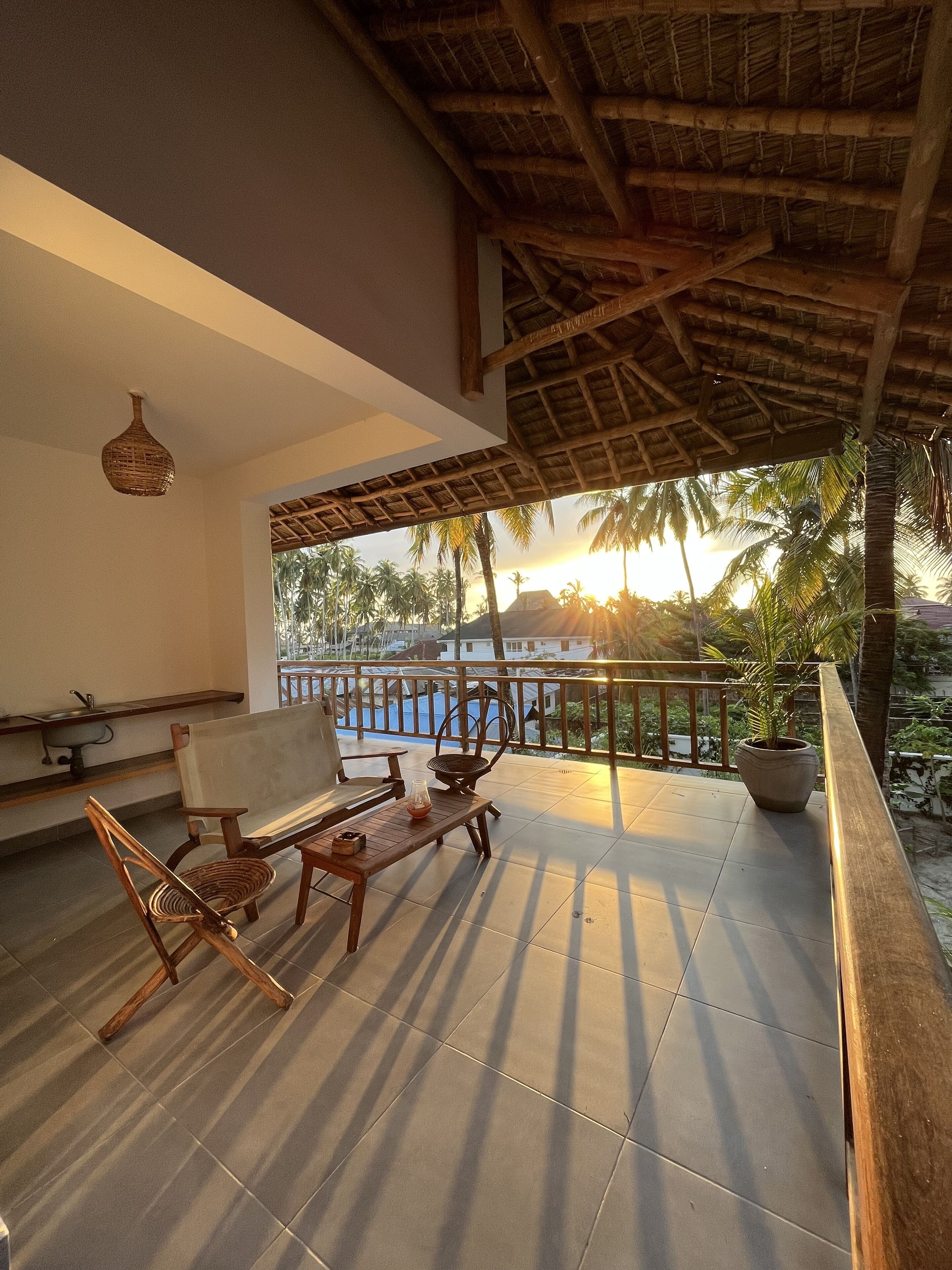 Un balcón con techo de paja, muebles de madera y vistas a palmeras y una piscina al atardecer.