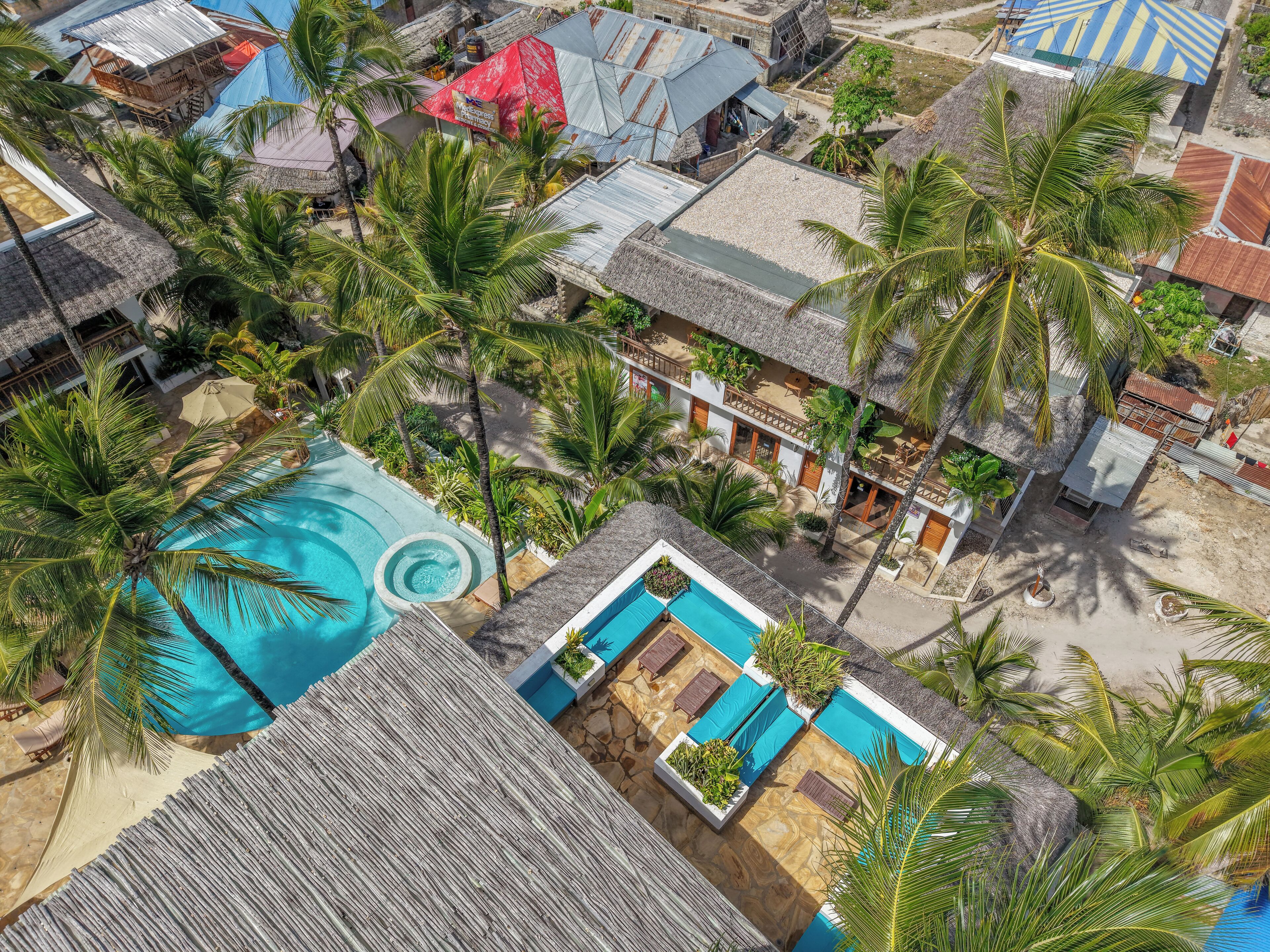 An aerial view of a resort with a swimming pool, palm trees, and thatched-roof buildings.