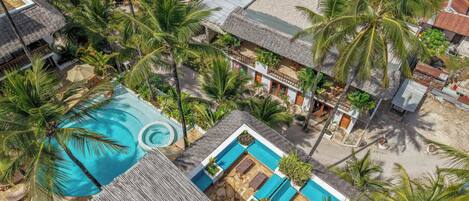 An aerial view of a resort with a swimming pool, palm trees, and thatched-roof buildings.