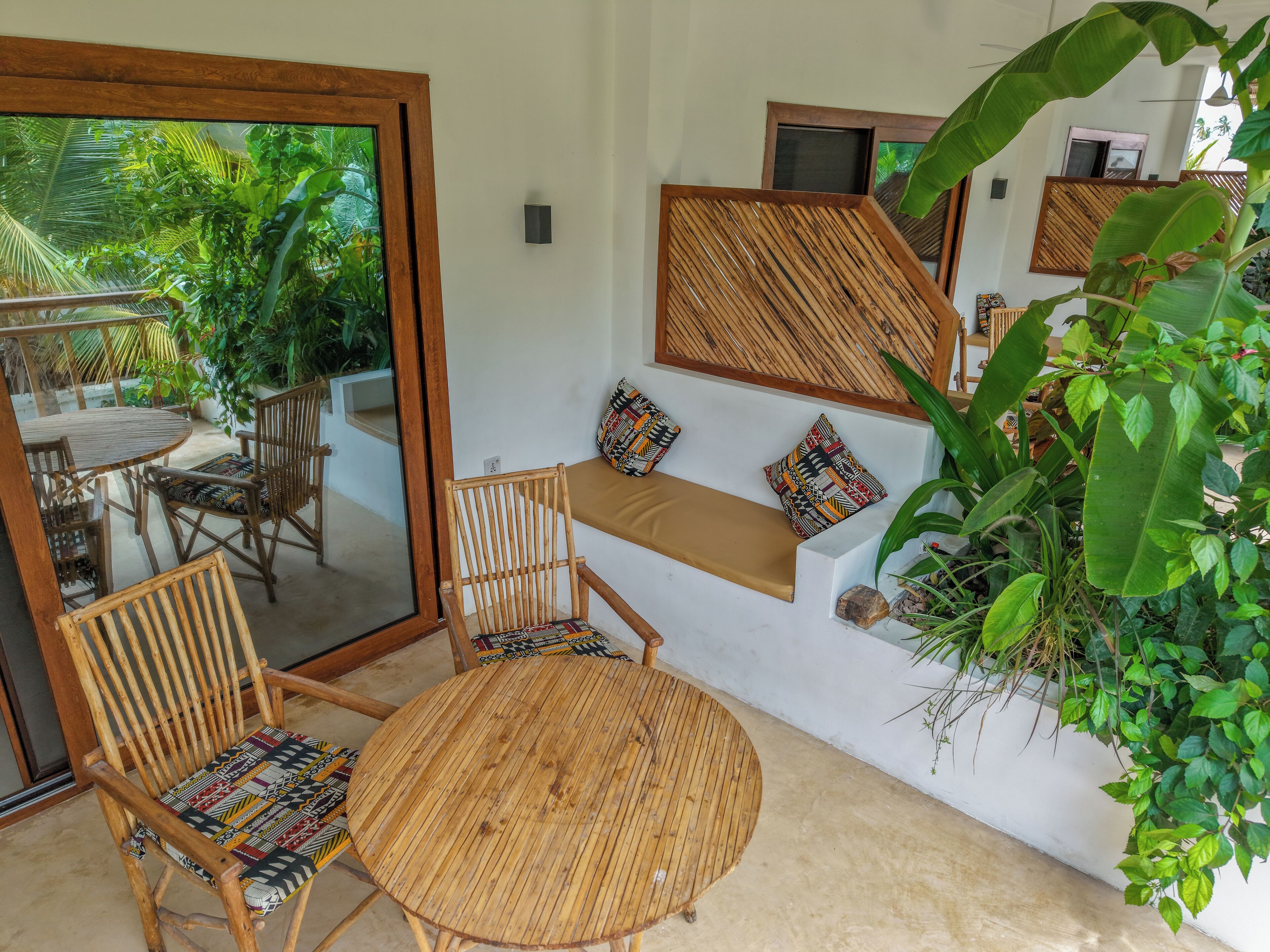 A room with a round wooden table, two wicker chairs, a bench with patterned cushions, and a large mirror.