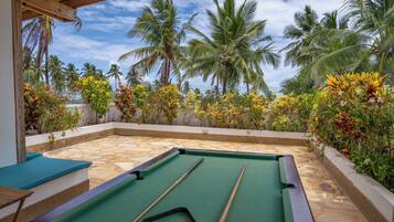 Área externa com piscina, teto de madeira, mesa de bilhar e plantas tropicais ao redor.