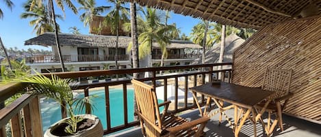 A balcony with a pool, wooden furniture, and a view of a resort with palm trees and buildings.