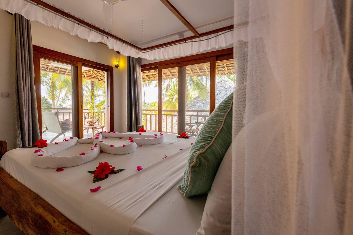 Ein Hotelzimmer mit einem Bett, das mit Rosenblättern dekoriert ist, einem grünen Kopfkissen und Blick auf Palmen durch große Fenster.