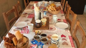 Une table à manger dressée avec du pain, du beurre, des confitures et des condiments, entourée de chaises en bois.