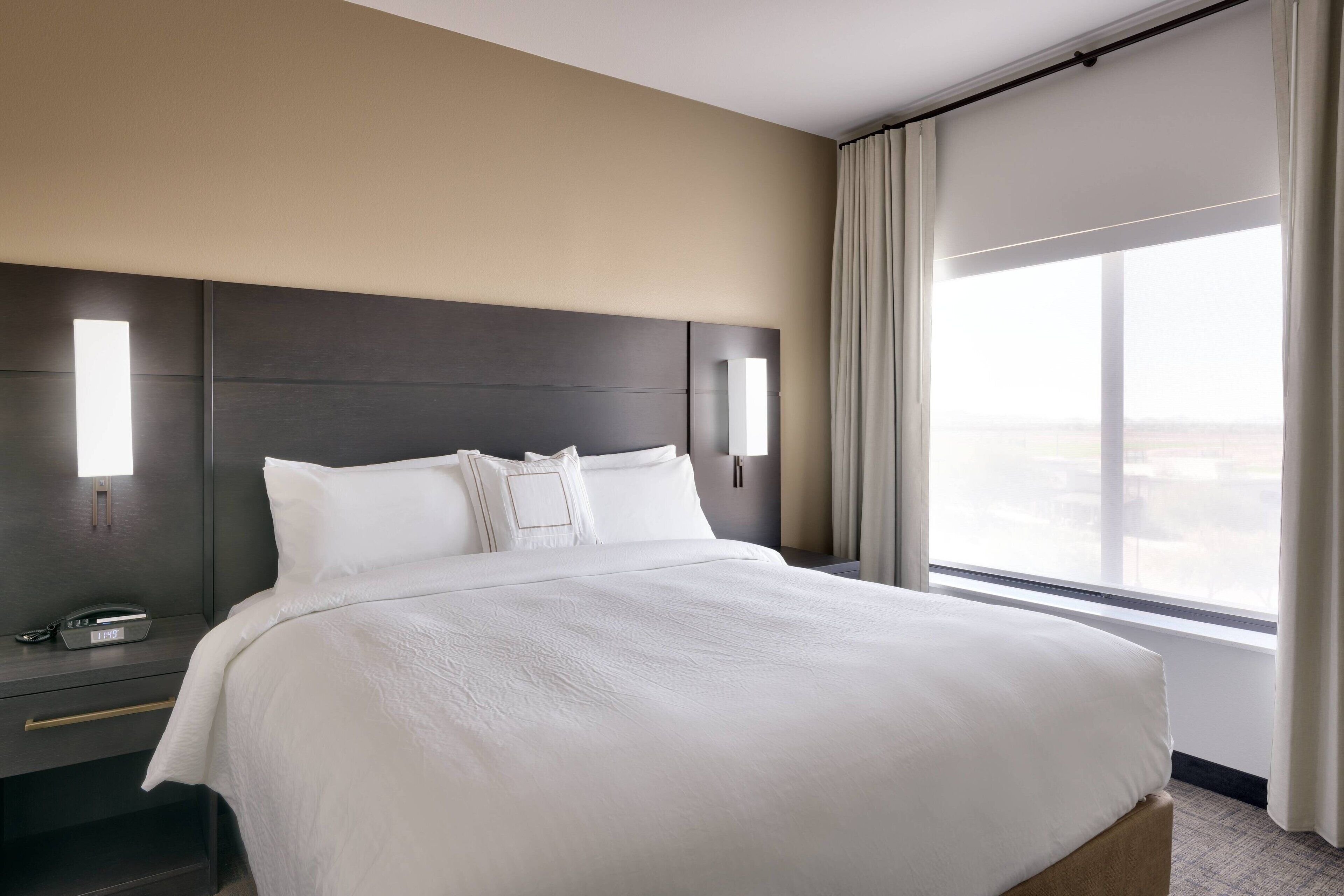 Quarto de hotel com uma cama grande, mesa de cabeceira com telefone e janela com cortinas.