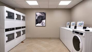 A laundry room with multiple washing machines and dryers, a tiled floor, and a wall-mounted abstract painting.