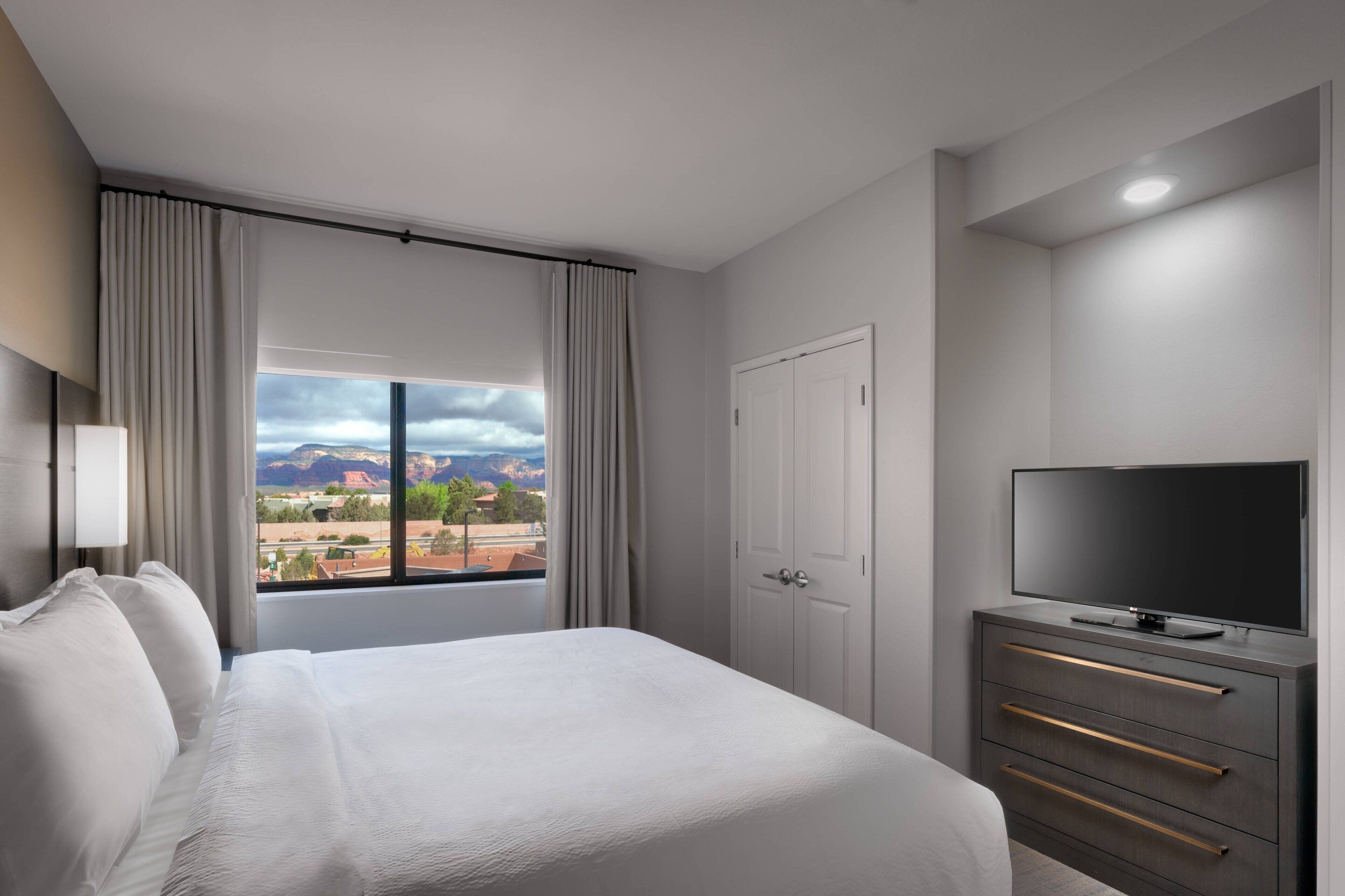 A hotel room with a large bed, a TV on a dresser, and a view of mountains through the window.