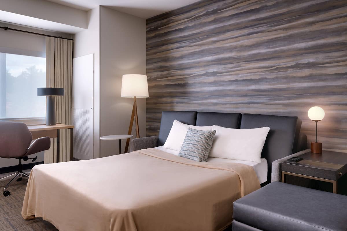 A hotel room with a large bed, a desk, a chair, a lamp, and a nightstand.