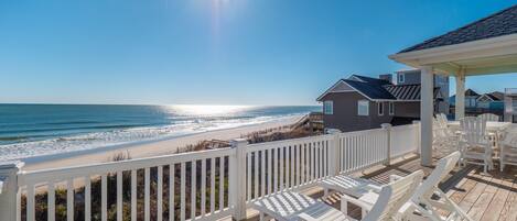 A wooden deck with white chairs, a view of the beach, and a house in the background.