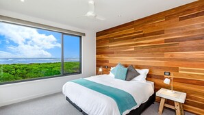 A bedroom with a large window, wooden wall paneling, a bed with pillows, a bedside table, and a ceiling fan.