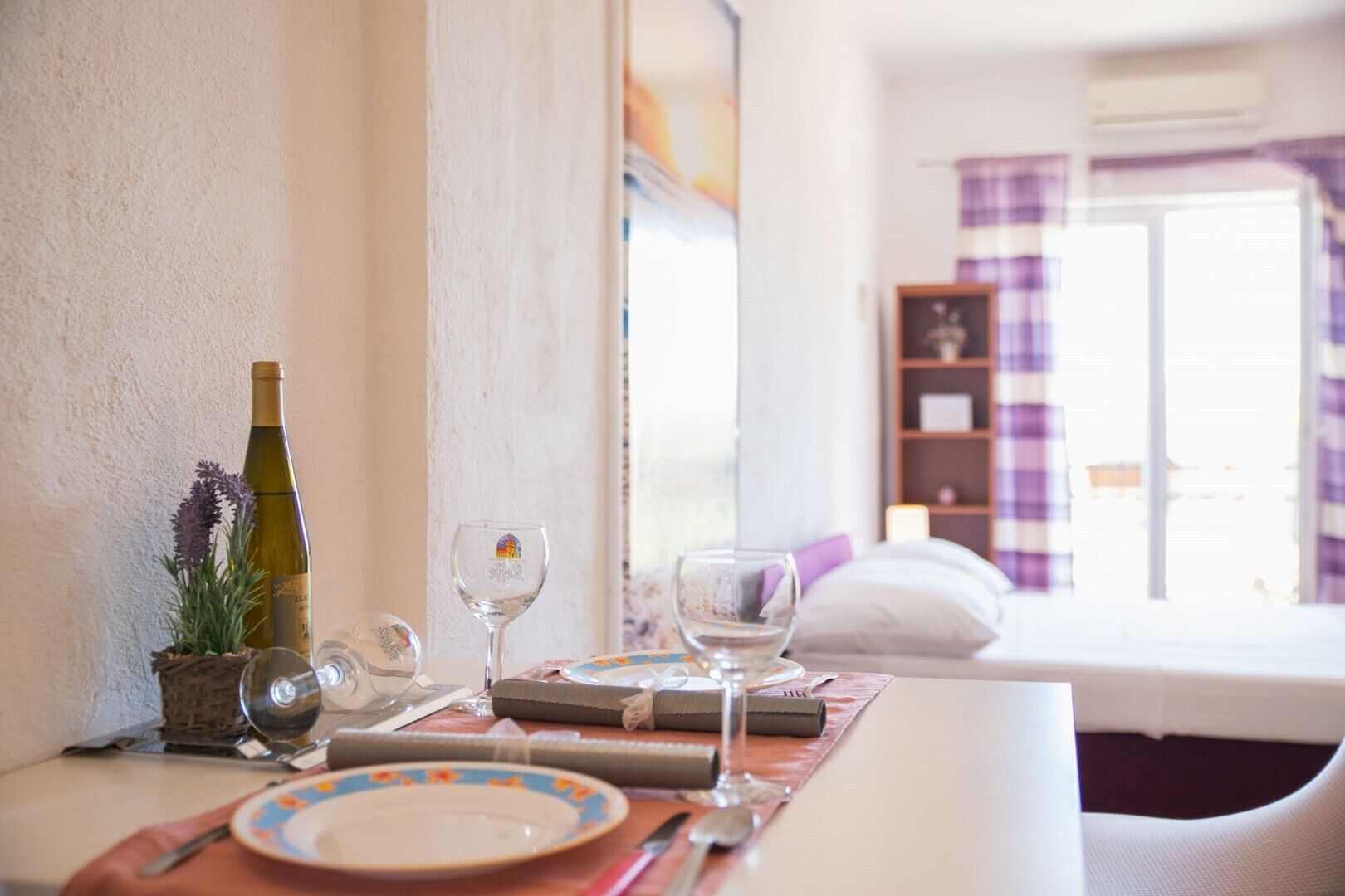 A neatly arranged dining table with a bottle of wine, glasses, and cutlery, set against a white wall with a window in the background.