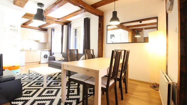 A dining area with a white table and dark wooden chairs, a black and white patterned rug, and wooden beams on the ceiling.