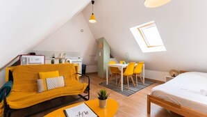 A compact, well-lit room with a yellow sofa, a bed, a dining area, and a small kitchen.