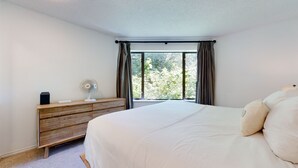 A bedroom with a bed, a wooden dresser, a standing fan, and a window with curtains. - Lovely, 2 bedroom / 2 bathroom, Villas Foxglove townhome short walk to the slope (Whistler)
