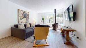 A modern living room with a grey sofa, a wooden rocking chair, a large painting, a TV, and a wood-burning stove. - Lovely, 2 bedroom / 2 bathroom, Villas Foxglove townhome short walk to the slope (Whistler)