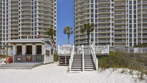 Un edificio frente a la playa con un pequeño quiosco, una escalera que lleva a un balcón y una palmera.