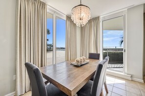 Uma sala de jantar com mesa de madeira, cadeiras cinzentas, um candelabro e vista para uma praia através de amplas janelas.