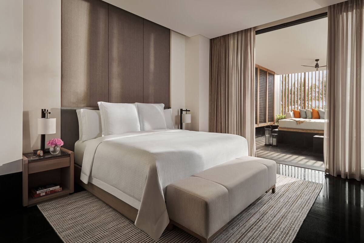 A modern hotel room with a large bed, a bedside table with a lamp, a small shelf with books, and a view of a balcony with a seating area.